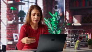 Mulher sentada em frente ao desktop olha com dúvidas para um cartão de crédito: será que ela é vítima de golpe digital?