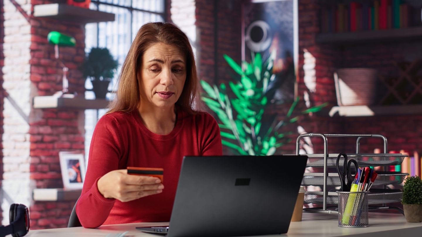 Mulher sentada em frente ao desktop olha com dúvidas para um cartão de crédito: será que ela é vítima de golpe digital?